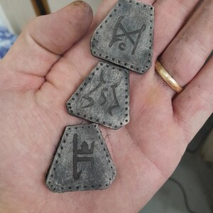 May include: Three trapezoidal, dark gray, textured patches with black stitched borders. Each patch features a unique, dark, etched-style symbol centered on the surface. The patches have a weathered, matte finish and are arranged in a vertical line.