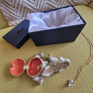 May include: A gray ceramic elephant trinket box with rhinestones, open to reveal a red interior. The box is sitting on a yellow surface next to a gold chain necklace with a small elephant pendant.