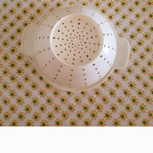 Peut inclure: Une passoire en plastique blanc avec de nombreux petits trous pour l'&eacute;gouttage. La passoire a deux poign&eacute;es et une base large. Le fond est un motif floral jaune et vert.