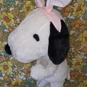 May include: A plush white Snoopy toy with black ears and a black nose, wearing a pink headband with white bunny ears. The toy is standing on its hind legs. The background is a floral pattern.