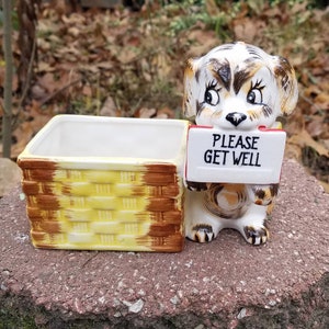 May include: A ceramic planter featuring a cartoon dog holding a sign that reads "PLEASE GET WELL." The dog is brown, white, and tan, with a basket-style planter in yellow and brown. The planter is on a stone surface.