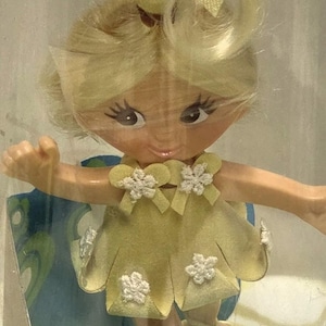 May include: A doll with blonde hair styled with a yellow bow, wearing a yellow dress with white flower accents. The doll is in a clear plastic case. The doll's name is "Spinderella Flatsy".