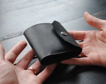 May include: A black leather wallet with a flap closure and a silver button. The wallet is folded in half and held in a hand.