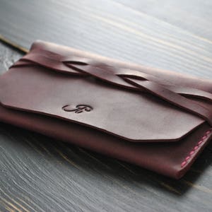 May include: A burgundy leather wallet with a braided leather strap and pink stitching. The wallet has a flap closure and a monogrammed initial.