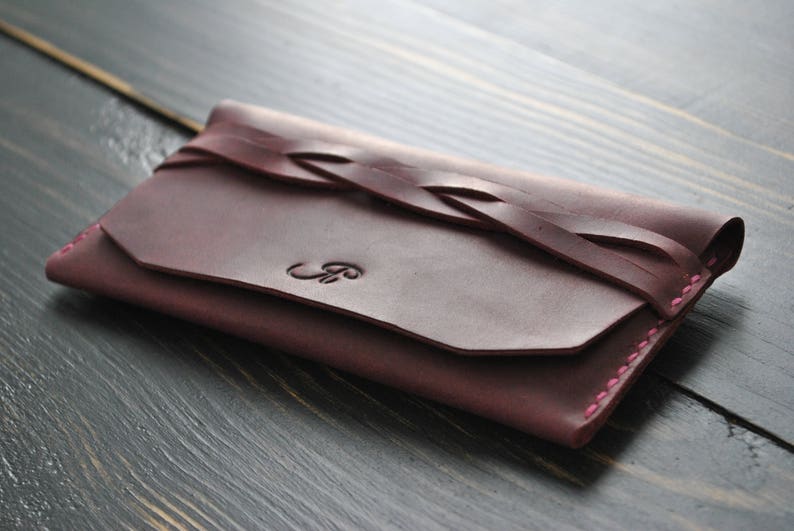 May include: A burgundy leather wallet with a braided leather strap and pink stitching. The wallet has a flap closure and a monogrammed initial.