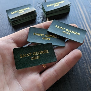 May include: Green rectangular business cards with gold lettering that reads "SAINT GEORGE ATELIER". Several stacks of cards are visible, with a few cards held in a hand. The cards have rounded corners and a textured surface.