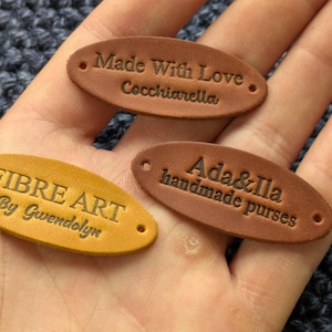 May include: Three oval-shaped leather tags with pre-punched holes on each end. One tag is yellow with the text FIBRE ART By Gwendolyn. Two tags are brown, one with the text Made With Love Cocchiarella and the other with Ada&Ila handmade purses.