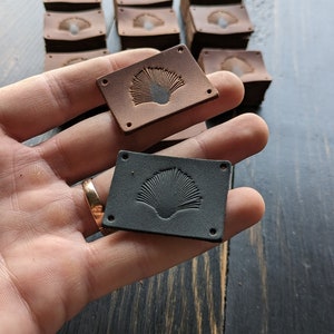 Könnte beinhalten: Zwei Leder-Etiketten mit einem dekorativen Design eines stilisierten Fächers. Ein Etikett ist braun und das andere ist schwarz.