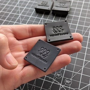 Op de afbeelding: Vier zwarte leren labels met elk vier gaten. De labels hebben een logo met drie gestileerde figuren met de tekst "VELVET LOOPS" eronder.