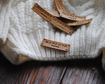 Può includere: Diverse etichette in pelle marrone con il testo "Nafanya Home" in rilievo. Le etichette sono sparse su un tessuto bianco a maglia.