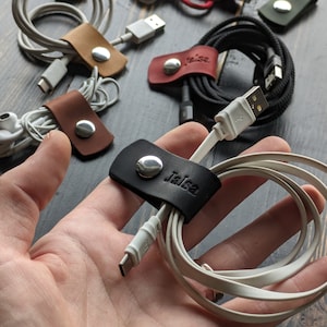 Puede incluir: Primer plano de un organizador de cables de cuero negro con el nombre "Jaisa" grabado en él. El organizador contiene un cable de carga blanco.
