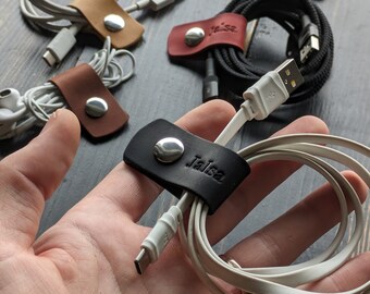 Könnte beinhalten: Nahaufnahme eines schwarzen Leder-Kabelorganizers mit dem eingeprägten Namen "Jaisa". Der Organizer hält ein weißes Ladekabel.