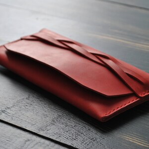 May include: A red leather wallet with a braided design. The wallet is closed and lying on a dark wood surface.