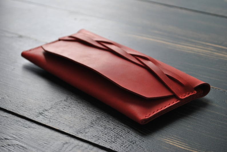 May include: A red leather wallet with a braided design. The wallet is closed and lying on a dark wood surface.