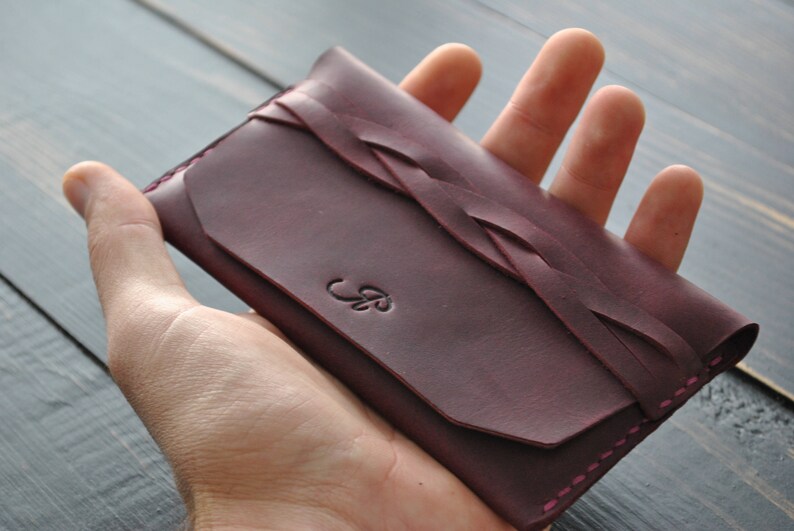 May include: A burgundy leather wallet with a braided design and a small leaf-shaped logo. The wallet is closed and has a pink stitch around the edges.