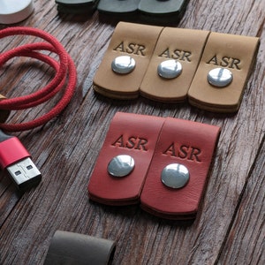 Puede incluir: Conjunto de seis organizadores de cables de cuero en verde, marrón y rojo. Cada organizador tiene un botón de metal plateado y las iniciales "ASR" grabadas en la parte delantera.