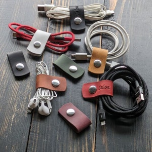 Puede incluir: Surtido de organizadores de cables de cuero en varios colores, incluyendo negro, blanco, marrón, verde, rojo y burdeos. Cada organizador tiene un botón de metal y algunos tienen texto personalizado, como "Jaisa".