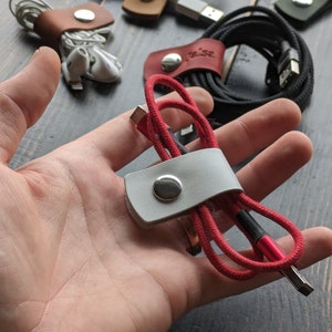 Puede incluir: Varios organizadores de cables de cuero en diferentes colores, incluyendo gris, marrón y rojo, con cierres de botón a presión plateados. Algunos organizadores sujetan cables enrollados, otros auriculares y cables de carga. Fondo de madera oscura.