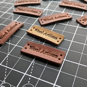 May include: Close-up of several brown leather labels with the text "Wool Artisan" embossed on them. The labels are arranged on a black cutting mat with white grid lines.