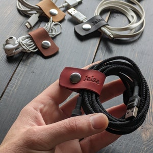 Puede incluir: Organizador de cables de cuero rojo con el nombre "Jaisa" grabado en él. El organizador contiene un cable de carga negro.