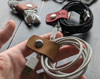 Könnte beinhalten: Nahaufnahme einer Hand, die einen braunen Leder-Kabelorganizer mit einem silbernen Knopf hält. Der Organizer hält ein weißes Ladekabel.