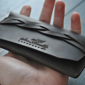 May include: A dark brown leather wallet with a braided design and the text "The Artfully" embossed on the front.