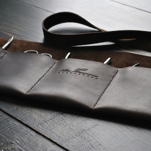 May include: A brown leather roll-up tool holder with a strap. The holder has multiple pockets for tools and is stitched with a decorative pattern. The holder has a brand name embossed on the front.