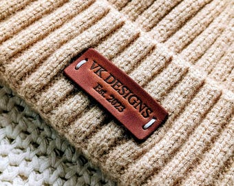 Può includere: Primo piano di un berretto lavorato a maglia a coste beige con un'etichetta in pelle marrone. L'etichetta recita "VK DESIGNS Est. 2023" in lettere bianche. Il berretto è appoggiato su una superficie bianca all'uncinetto.