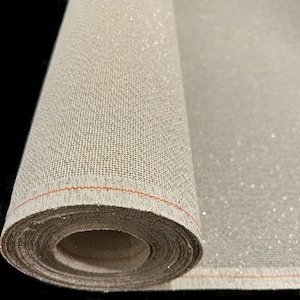 May include: A rolled-up roll of beige fabric with a subtle sparkle. The fabric has a woven texture and a thin orange line along the edge. The material appears to be a decorative textile, possibly for crafts or home decor.