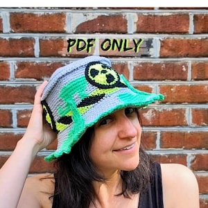 May include: A crocheted hat in shades of gray, green, black, and yellow, featuring a radiation symbol design. The hat has a brim and is labeled "PDF ONLY" in the image.