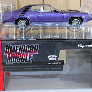 May include: A purple die-cast Plymouth model car in a clear plastic blister pack sitting on a black and red American Muscle box. The box features the text American Muscle, Plymouth, Looney Tunes, and Limited Edition with white and red accents.