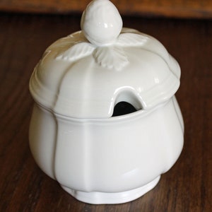 May include: A white ceramic sugar bowl with a lid and a small handle. The lid has a leaf design and a small knob on top. The bowl has a round base and a curved body. The sugar bowl is sitting on a wooden surface.