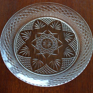 May include: Clear glass plate with intricate diamond and starburst patterns. The plate features a detailed design around the rim and a central starburst motif. This decorative plate is suitable for serving or display.