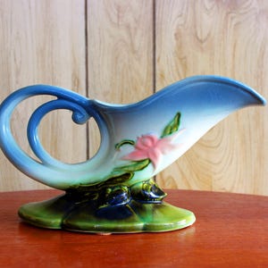 May include: A vintage ceramic pitcher with a blue and white gradient, featuring a decorative swirl handle. The pitcher is adorned with a pink floral design and rests on a green base. A unique piece for display or serving.