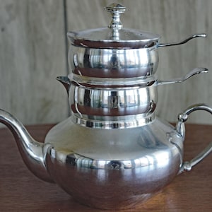 May include: A polished silver-tone metal tea set consisting of three stacked components. The base is a wide teapot with a curved spout and handle, topped by two smaller, cylindrical containers, each featuring a small side handle and a lid.