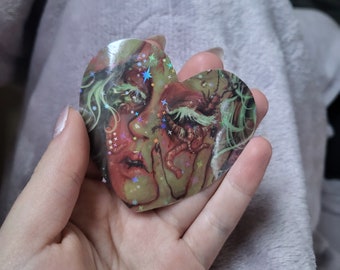 Puede incluir: Una pegatina en forma de corazón con una ilustración surrealista de un rostro con cabello verde, tonos de piel rojos y detalles en forma de gusano. La superficie tiene un acabado holográfico con pequeños reflejos de luz en forma de estrella.