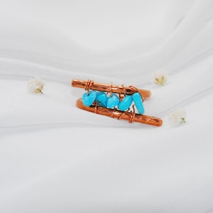 May include: A copper wire ring with turquoise stones wrapped around it. The ring is on a white fabric background.
