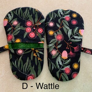 May include: A pair of black oven mitts with a floral pattern of pink and yellow flowers on a dark green leafy background. The mitts have a green fabric lining and a black loop handle. The text "D - Wattle" is printed below the mitts.