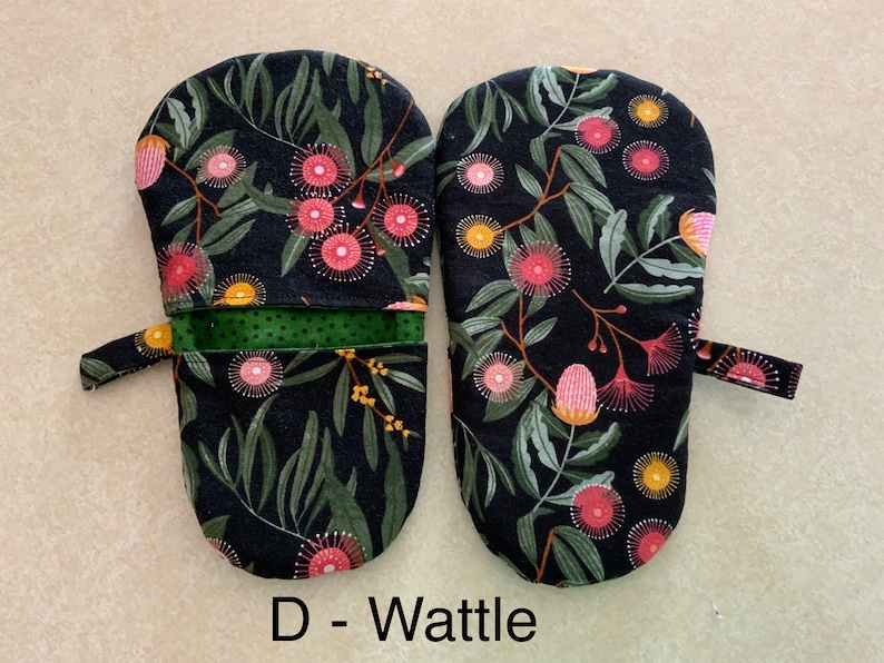 May include: A pair of black oven mitts with a floral pattern of pink and yellow flowers on a dark green leafy background. The mitts have a green fabric lining and a black loop handle. The text "D - Wattle" is printed below the mitts.
