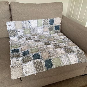 May include: A patchwork quilt in white, grey, and green, featuring patterns like dots, arrows, and animal motifs. The quilt is displayed on a beige sofa, highlighting its soft, textured design. The quilt's edges have a frayed, rag-style finish, adding to its charm.