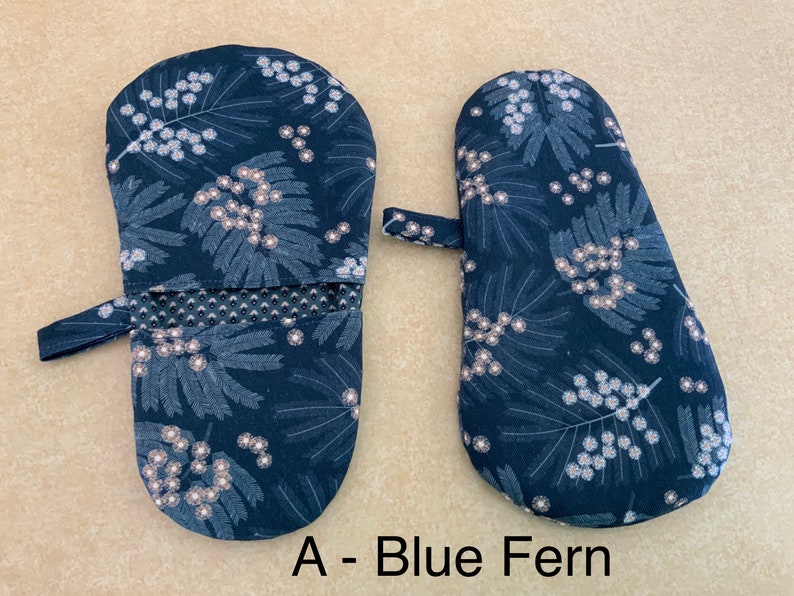 May include: A pair of blue oven mitts with a white floral pattern. The mitts have a hanging loop and a heat-resistant lining. The text "A - Blue Fern" is printed below the mitts.