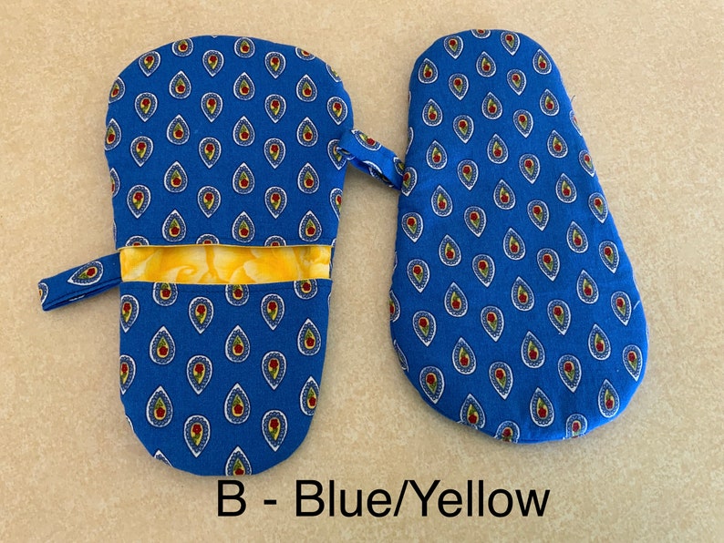 May include: A pair of blue oven mitts with a yellow and white paisley pattern. The mitts have a hanging loop and a pocket on the back. The text "B - Blue/Yellow" is printed below the mitts.
