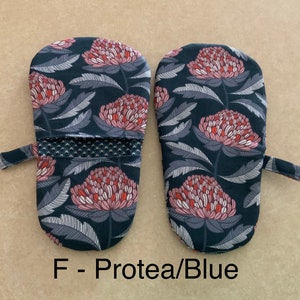 May include: A pair of blue oven mitts with a floral pattern featuring pink and red protea flowers. The mitts have a black and white striped trim and a hanging loop.  F - Protea/Blue