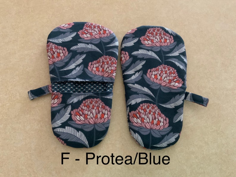 May include: A pair of blue oven mitts with a floral pattern featuring pink and red protea flowers. The mitts have a black and white striped trim and a hanging loop.  F - Protea/Blue