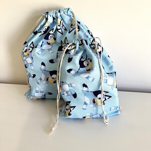 May include: Two light blue drawstring bags featuring a repeating cartoon dog pattern. The bags have white drawstrings and are made of fabric. One bag is larger than the other. The bags are suitable for storage.