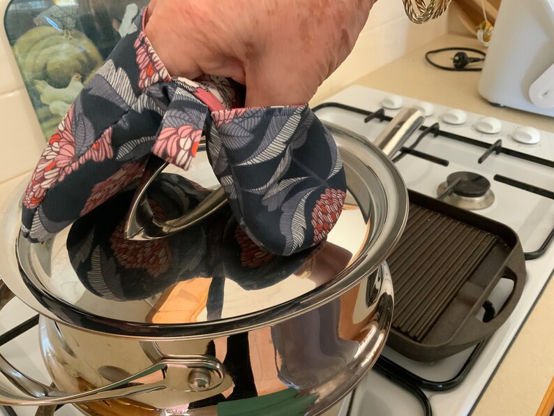 May include: A person's hand wearing a floral print oven mitt is holding the lid of a stainless steel pot on a hob. A black cast iron grill pan is on the hob to the right of the pot.