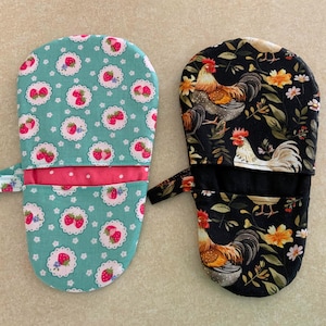 May include: Two oven mitts. One is teal with a strawberry pattern, the other is black with a rooster and floral design. Both have a loop for hanging and a red polka dot accent.