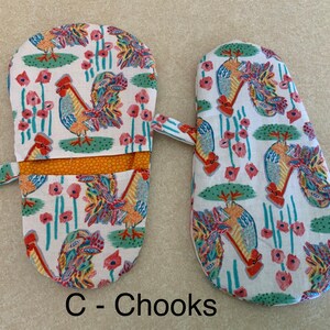 May include: A pair of oven mitts with a rooster and flower pattern. The mitts are white with a yellow, orange, red, blue, and green rooster design. The mitts have a white fabric strap and a yellow and orange lining. The text "C - Chooks" is printed below the mitts.