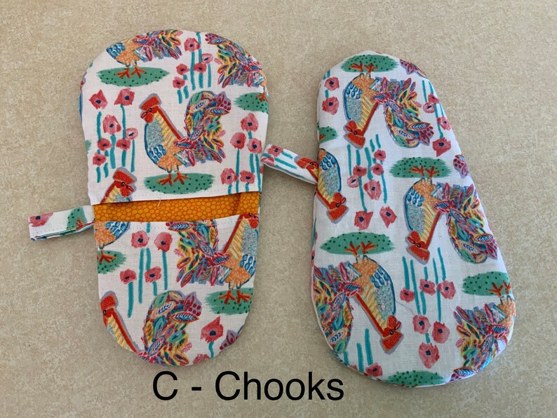 May include: A pair of oven mitts with a rooster and flower pattern. The mitts are white with a yellow, orange, red, blue, and green rooster design. The mitts have a white fabric strap and a yellow and orange lining. The text "C - Chooks" is printed below the mitts.