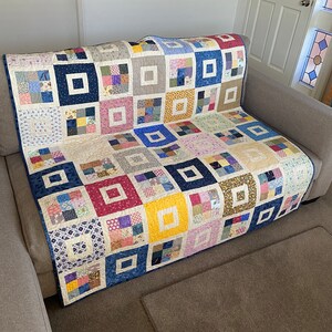 May include: A patchwork quilt with a variety of colourful squares and a white background. The quilt is folded and draped over a sofa.